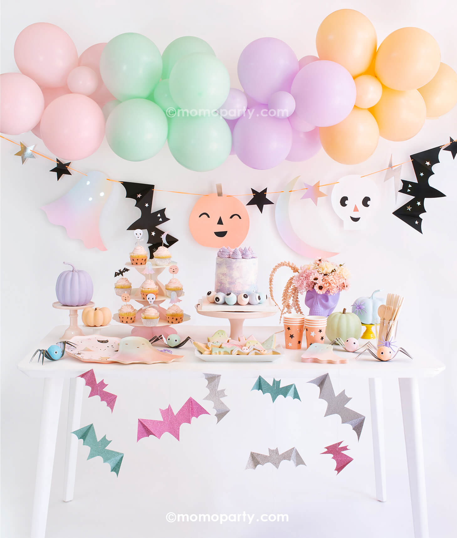Momo Party Pastel Halloween Box Kit. Set up with Meri Meri Pastel Halloween Dinner Plates, Pastel Ombre Halloween Ghost Plates, Pastel Halloween Star Pattern Cups, Pastel Ombre Halloween Ghost Napkins, Wooden Cutlery, Iridescent Party Straws, Pastel Halloween Cupcake Kit  with 24 toppers, cake with eyeballs in the pink cake stand, skull head with flowers, spider surprise balls on the table. Pastel Halloween Glitter Hanging Bats, Pastel Balloon Cloud and Pastel Halloween Garland Set as decoration 