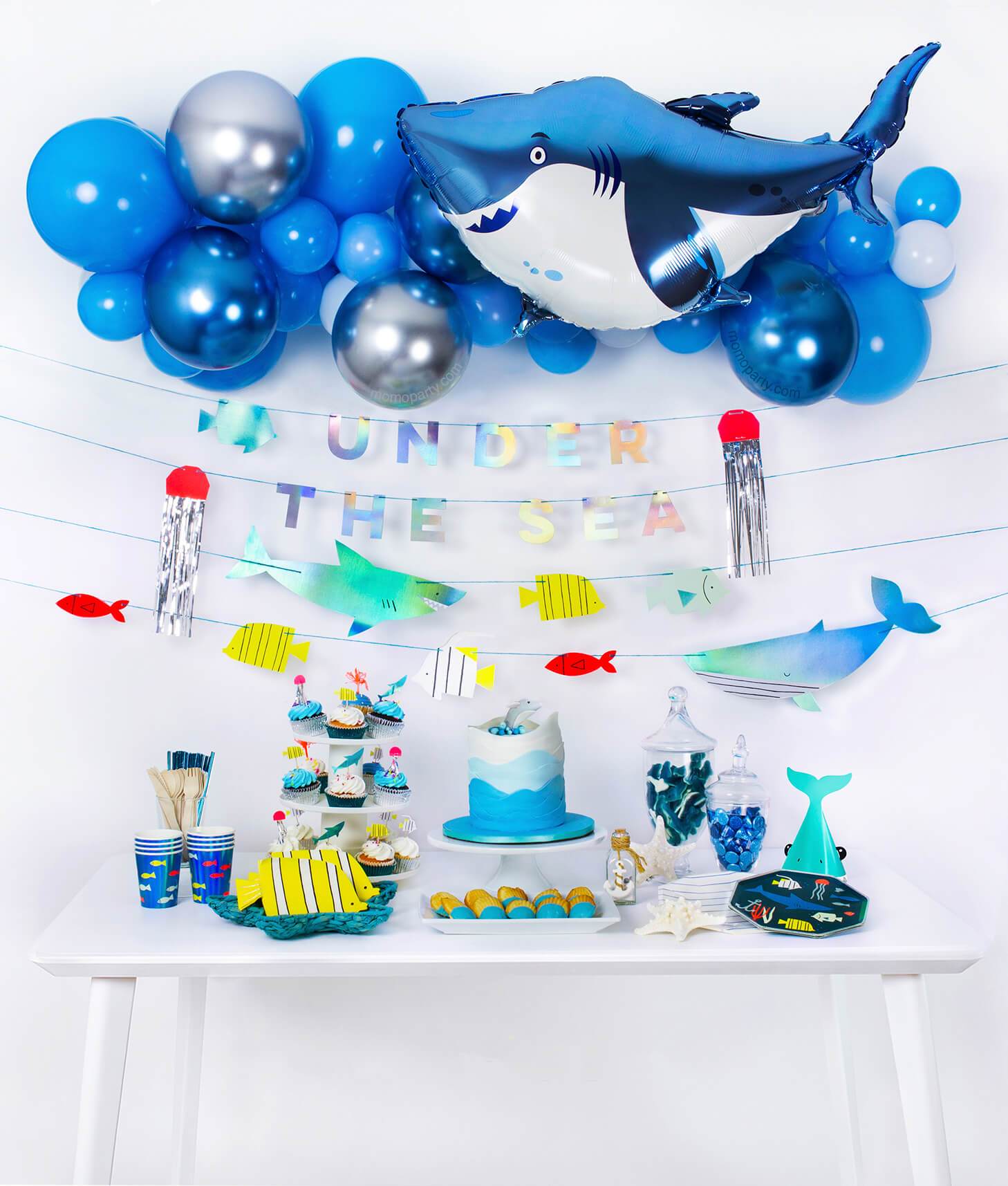 Momo Party Box of under the sea shark themed birthday party idea with balloon garland, shark foil balloon, Under the sea garland as decoration and fish graphic paper cups, fish die-cut napkins, shark party hats, plates and cupcakes as table set up