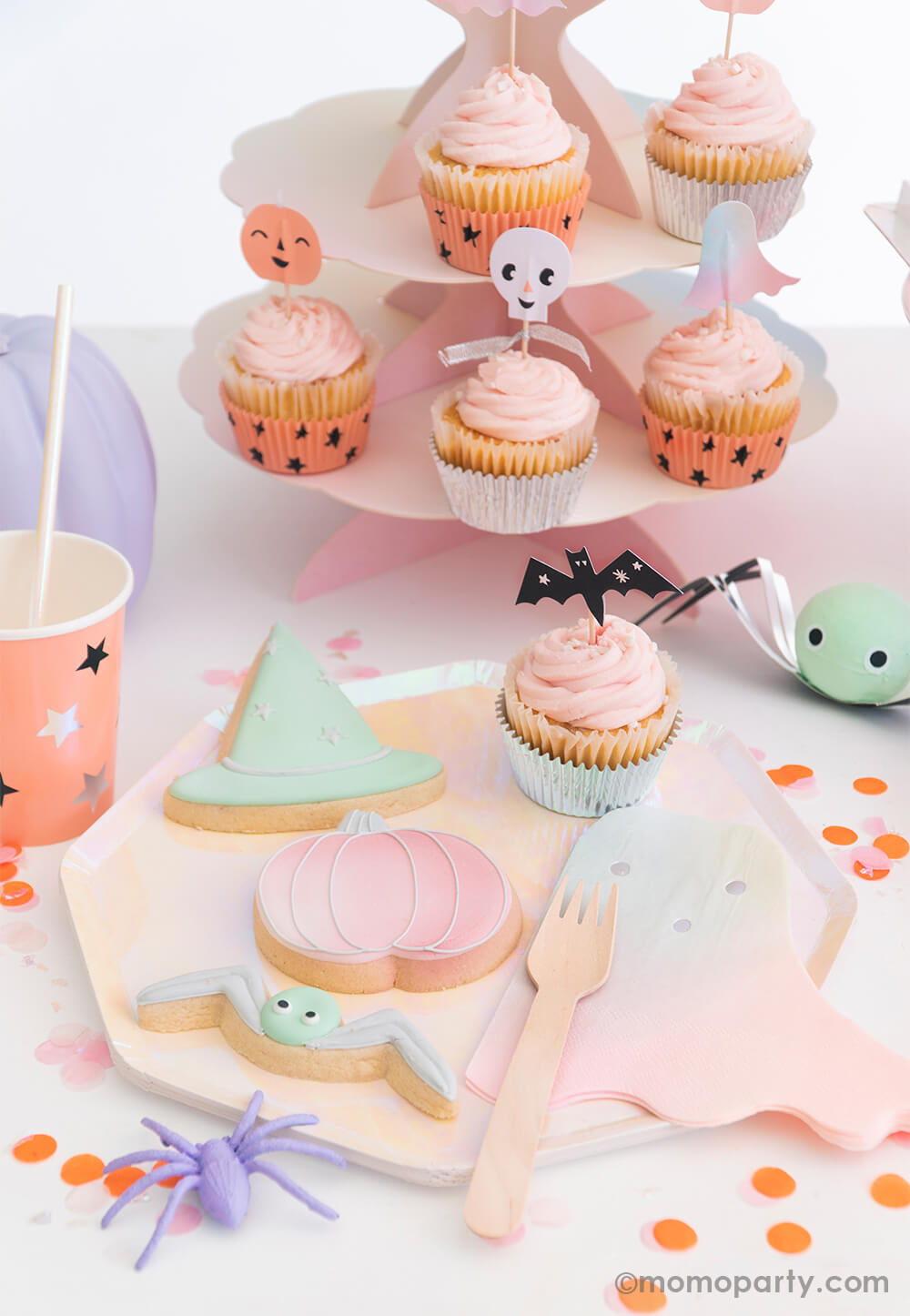 Pastel halloween party by momo party. Table set up with spider, pumpkin and witch hat shaped cookies, and a bat topper on a cupcake, Pastel Halloween ghost napkin and wooden fork on a Meri Meri iridescent dinner plate. A Pastel Halloween Star Pattern Cup and mint spider surprise ball and confetti around the plate. cupcakes decorated with Pastel Halloween Cupcake Kit with 6 terrific designs of toppers - skulls, stars, pumpkins, ghosts on a pastel pink cake stand, Halloween party supplies for pink halloween