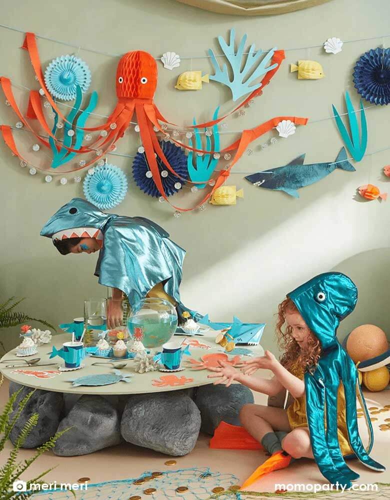 A festive party table filled with Momo Party's sea collection by Meri Meri, featuring tableware in sea creature designs including turtles, octopus, lobsters, sharks. On the wall is a large party garland set with honeycomb octopus and blue paper fan decorations, transforming the room to an underwater space. All makes great inspiration to kid's under the sea themed parties.