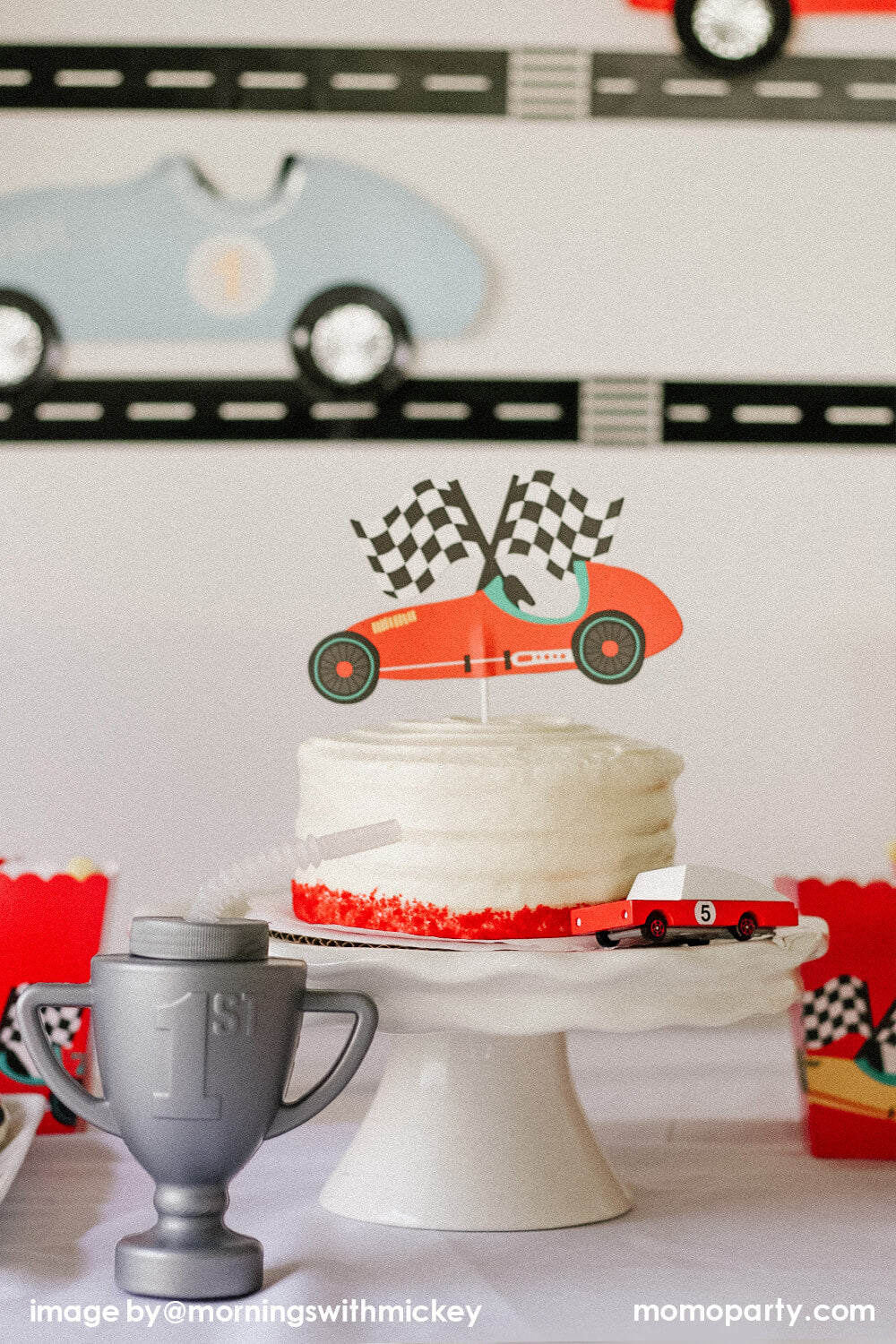 Race Car-themed Birthday party table close-up of, a Buttercream cake on a white cake stand decorated with Merrilulu Custom Race Car Cake Topper, a CANDYCAR RED RACER toy on the side, and a RACE CAR TROPHY CUP next to it. This modern Race Car Birthday party hosted by Momo Party with Mornings-with-Mickey is so fun for kids who love Disney Cars, Race Car and “two fast” 2nd birthday