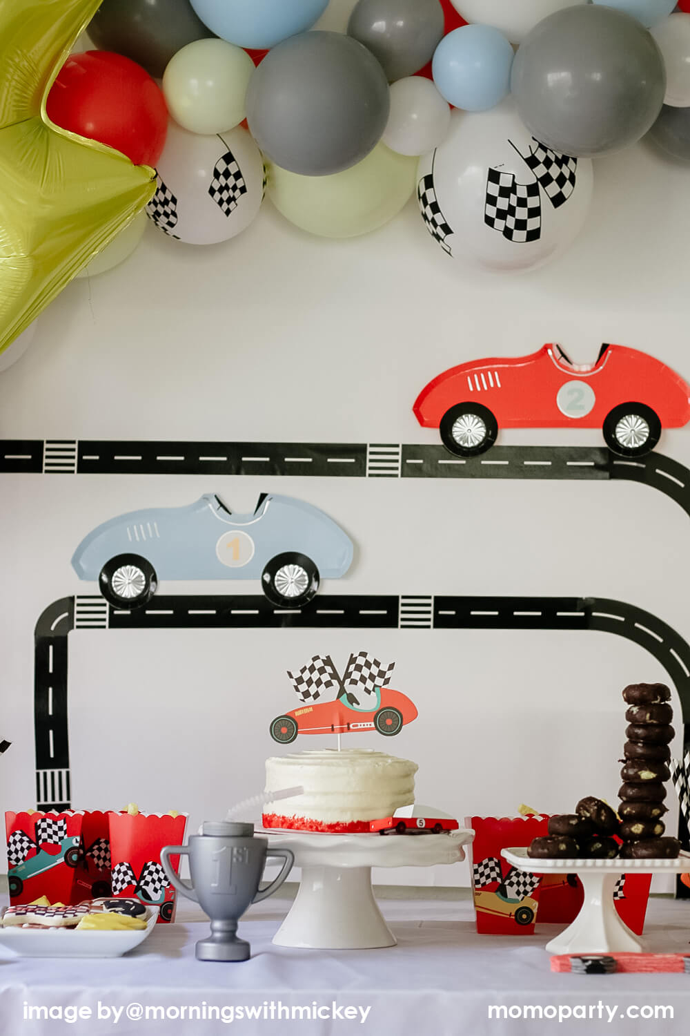 Modern disney race car party, decorated with Road Tape on the wall with Meri Meri Race Car Die-cut Paper Plates, modern pastel themed balloon cloud mix with flag latex balloon and lighting bolt foil balloon, race car topper on a buttercream cake, trophy sipper, mini donuts as tire on a cake stand, red popcorn cups with race car stickers next to the main cake. A modern Car themed Birthday Party at home by Mornings-with-Mickey