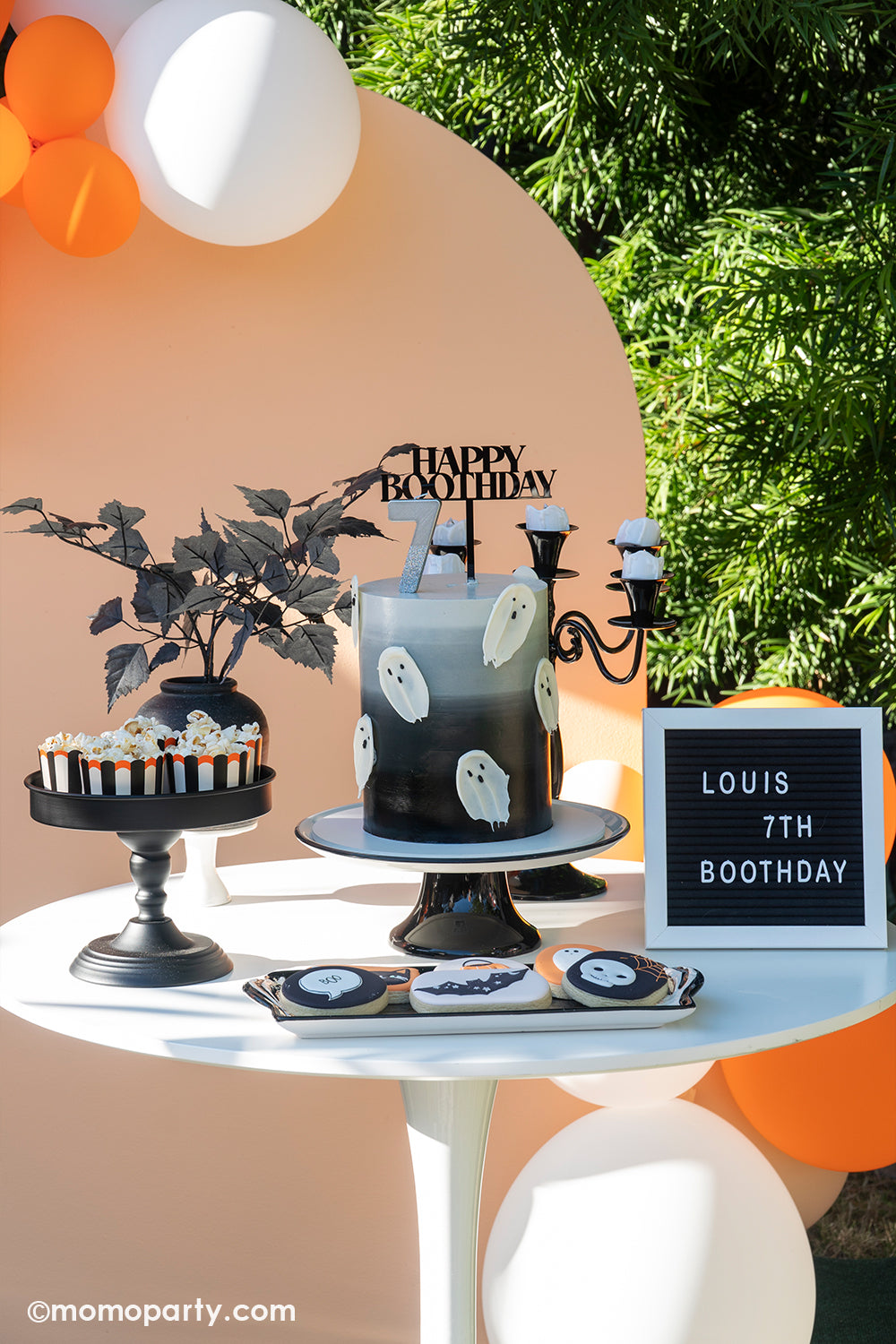 A gorgeous dessert table for a kid's Halloween themed birthday featuring an black to grey ombre buttercream cake decorated with ghost shaped melted chocolate, a range of Halloween themed sugar cookies in the designs of bats, skulls, black cats, ghosts, plus some Halloween themed food cups featuring black and white stripes with orange scallop edge filled with popcorn for kids' snacks
