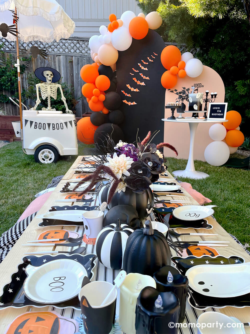 A spooky fun kid-friendly Halloween party set up featuring classic orange and black color themed party supplies including My Mind's Eye bat shaped paper decorations in orange on the backdrop which was behind an adorable kid low picnic table filled with fun Halloween themed party tableware including ghost shaped plates, jack o' lantern pumpkin bucket shaped napkins with black and white pumpkins, candles, skulls and Halloween themed floral arrangement as the centerpiece.