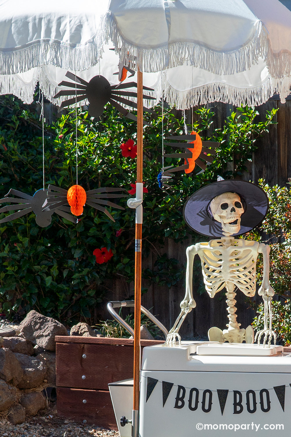 A spooky fun Halloween party set up with a paleta cart decorated with My mind's Eye Vintage Halloween Boo banner with Meri Meri's honeycombs in three different colors of orange, navy and mint greenhung from the boho themed umbrella, standing next to the paleta cart was a full sized posable skeleton wearing a witch hat, creating a spooky vibe for a Halloween celebration.