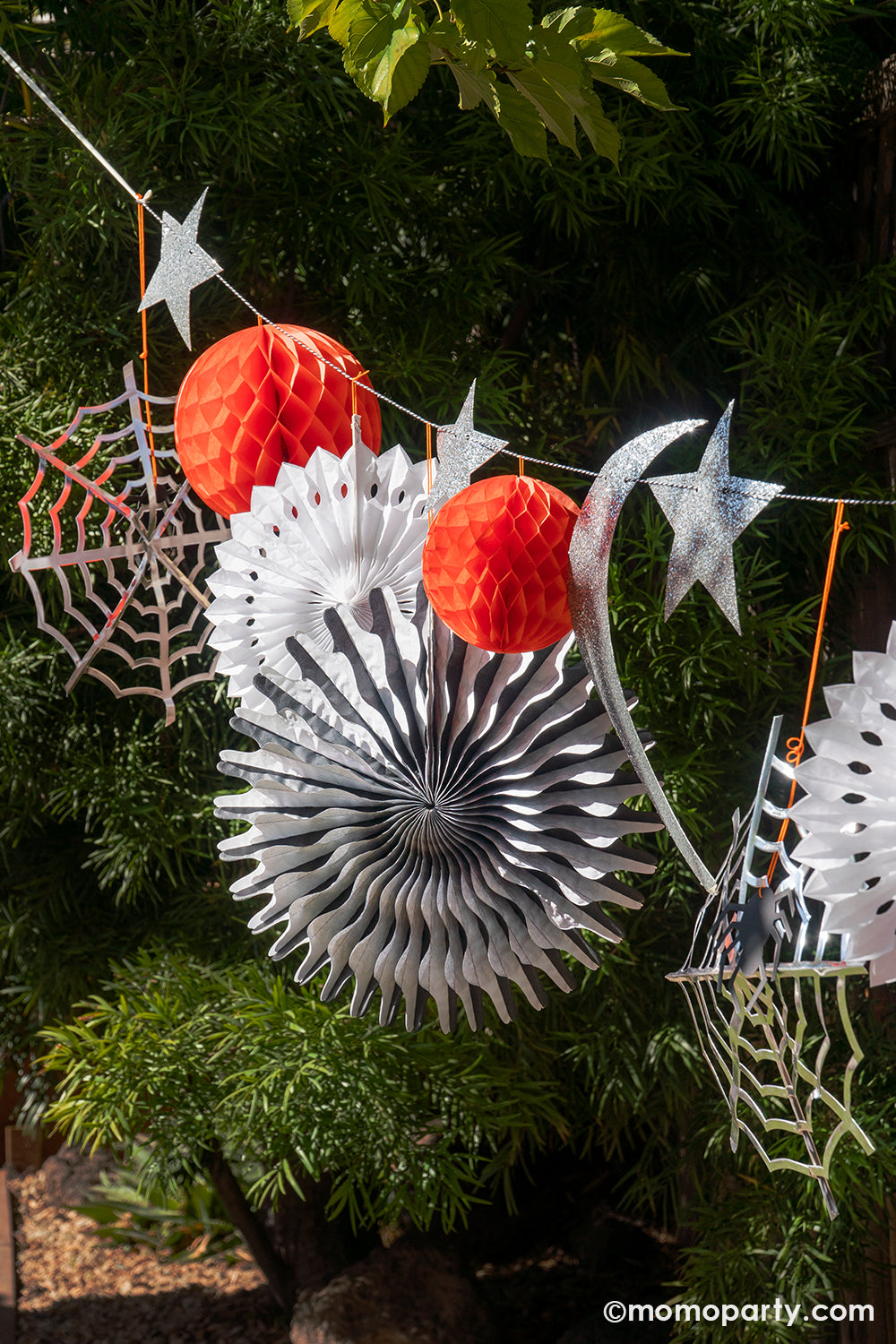 A close up of Meri Meri's Halloween honeycomb garland set in a backyard setting for a Halloween party. This set of garland includes a combination of stars, cobwebs, spiders, pinwheels, a crescent moon, honeycomb balls and lots of shimmering glitter, all in all makes an amazing decoration for a spooky fun Halloween celebration.