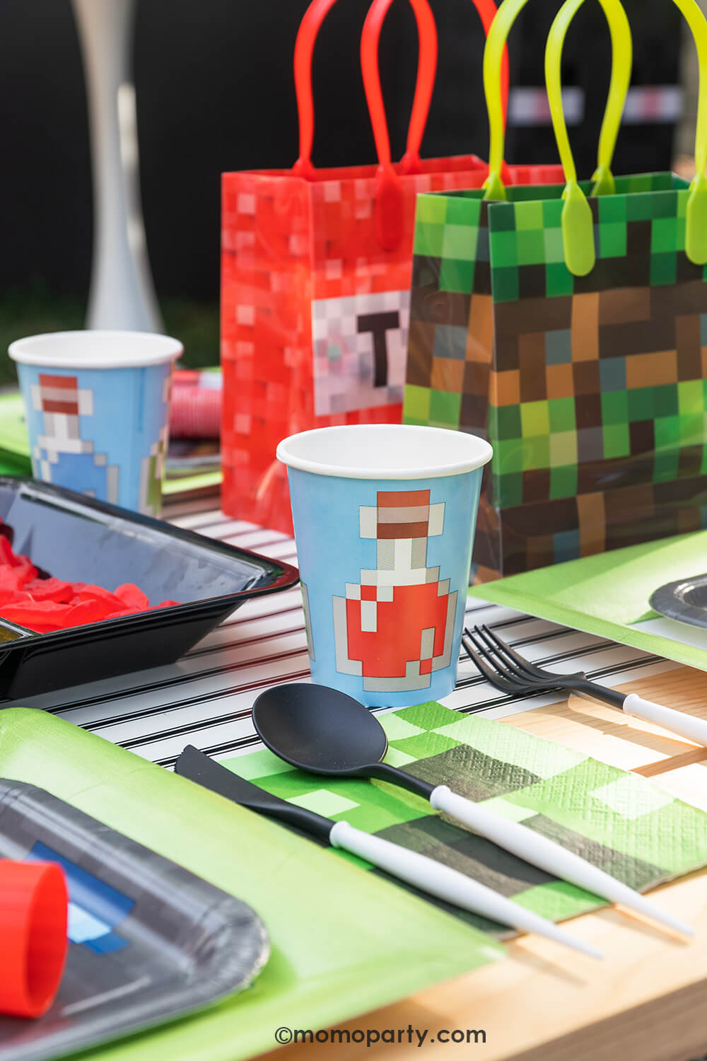 Minecraft themed Birthday Party Ideas by Momo Party. Featuring blue Minecraft potion paper cup with image of a potion bottle print, Minecraft gray side plate and modern sophisticated  large lime green dinner plates,  beautiful black and white cutlery sets, an pixel TNT theme goodie bags, all on the black striped table runner of the kids picnic table. 