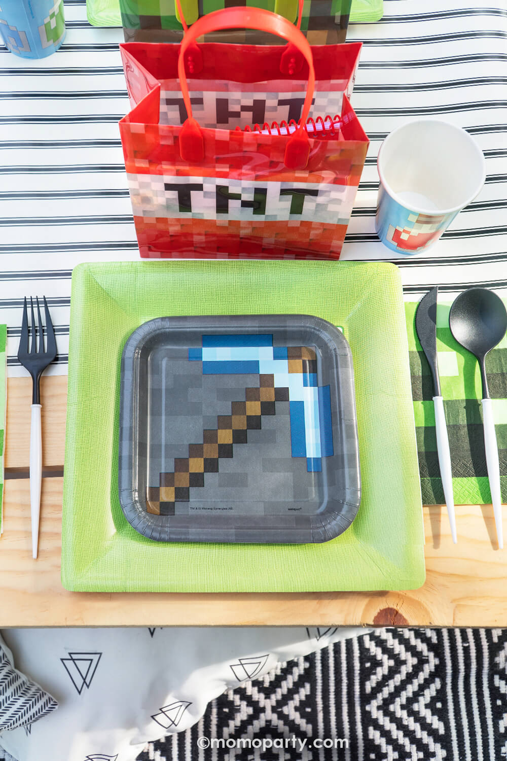 Top view of Minecraft themed Birthday Party Ideas by Momo Party. Featuring a fun Minecraft TNT Twist Poppers layered with Minecraft gray side plate and modern sophisticated  large lime green dinner plates, blue Minecraft potion paper cups,  beautiful black and white cutlery sets, an pixel TNT theme goodie bags, all on the black striped table runner of the kids picnic table. 