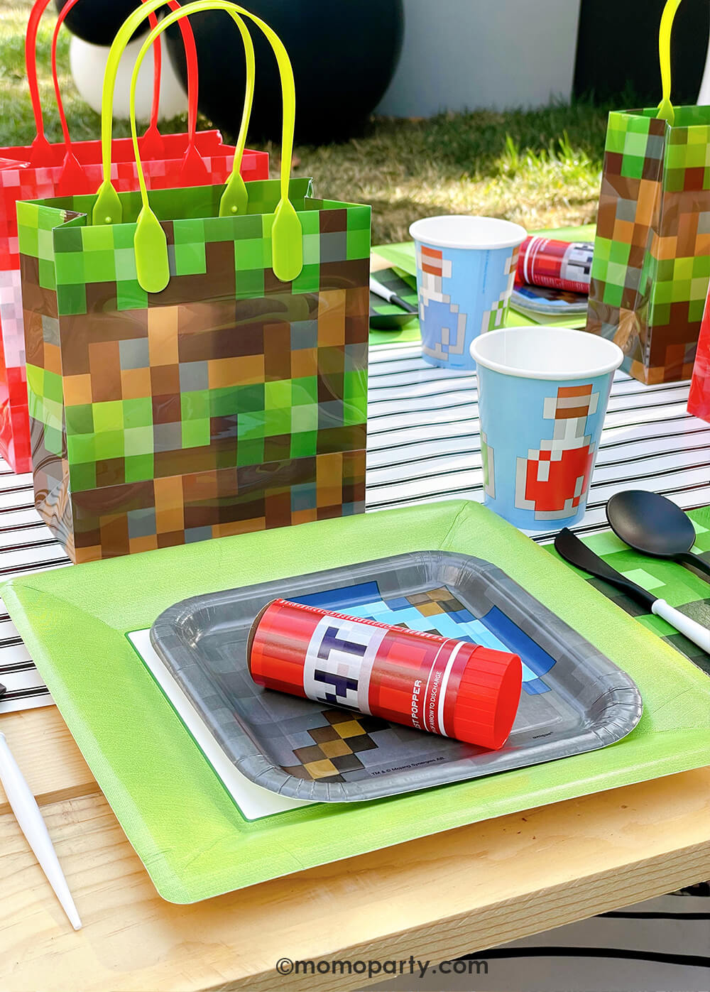 Minecraft themed Birthday Party Ideas by Momo Party. Featuring a fun Minecraft TNT Twist Poppers layered with Minecraft gray side plate and modern sophisticated  large lime green dinner plates, blue Minecraft potion paper cups,  beautiful black and white cutlery sets, an pixel TNT theme goodie bags, all on the black striped table runner of the kids picnic table. 
