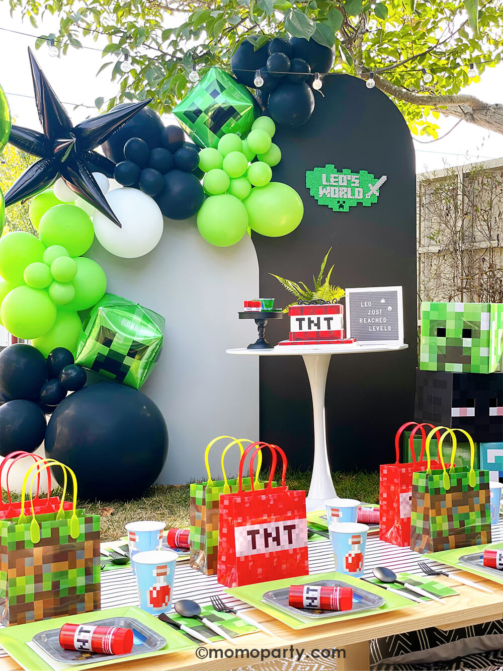 Kid's Minecraft themed Birthday Outdoor Party Ideas by Momo Party. Featuring Backdrop board in black and grey, decorated with minecraft themed color balloons in green, black, white, and creeper foil balloon, black starburst foil balloons. A TNT cake with letter board on the white round dessert table . In the front there is a kids picnic table filled with minecraft themed party plates with Minecraft Twist Poppers on top, cups, Creeper napkins and pixel TNT theme goodie bags on the black stripe table runner.