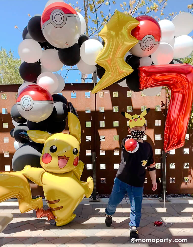 A boy wearing pikachu hat, holding a pokemon ball on his hand, standing on his Pokemon themed birthday Party. Decorated with a Black and white latex balloon garland, mix with Pokemon Pokeball Orbz Foil Balloon and Lightning Bolt Foil Balloon. A anagram 57" Pokemon Pikachu Airwalker Foil Balloon stand on the left, and a Large Number 7 Red Foil Mylar Balloon on the right side. A modern cute backdrop for a pokemon themed birthday party