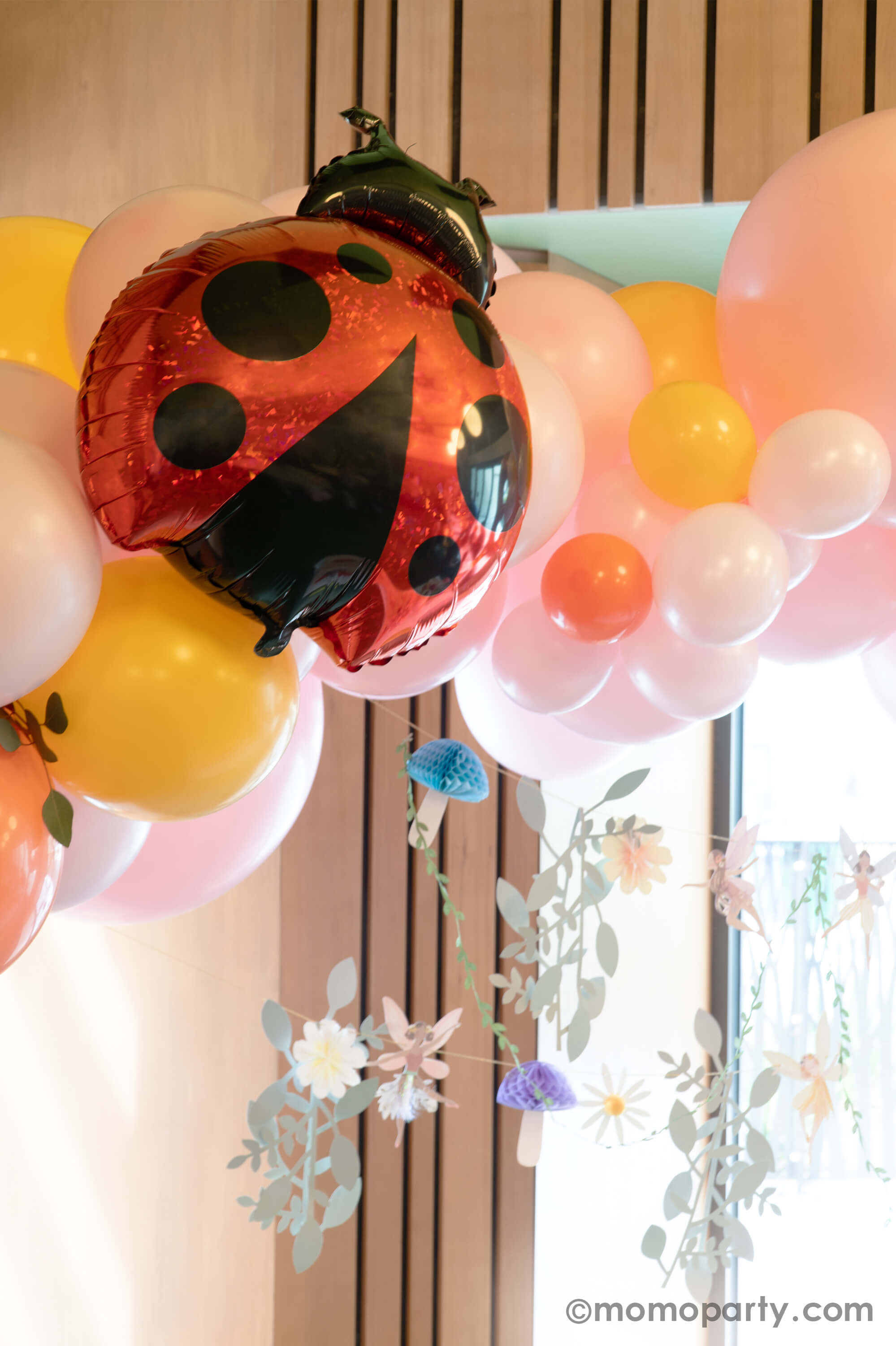 A spring fairy themed birthday party decoration featuring Momo Party's 27" ladybug shaped holographic foil balloon by Betallic Balloons on a spring inspired balloon garland in pastel pink, goldenrod, coral colors, with Momo Party's 6ft fairy garland by Meri Meri hung below it. All creates a beautiful decoration set up for a girl's birthday party in spring.