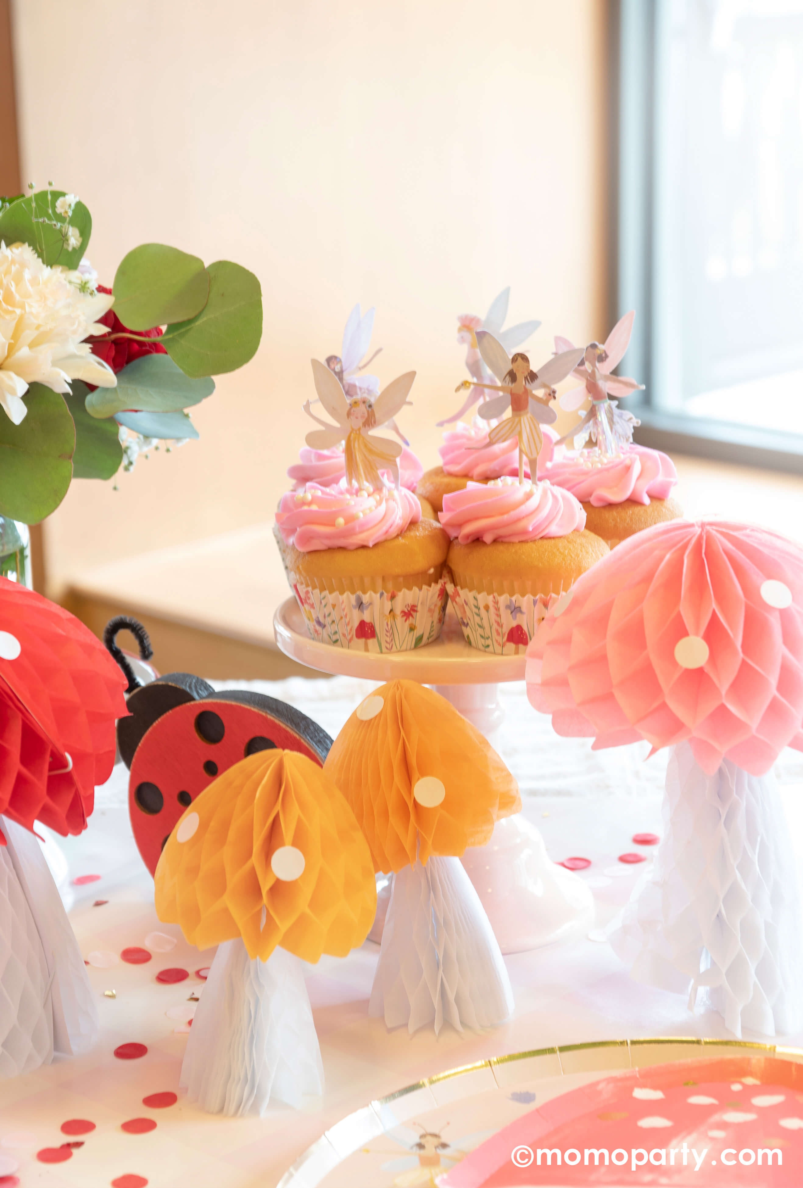 A spring fairy themed birthday party table featuring Momo Party's fairy mushroom shaped plate by Meri Meri, ladybug shaped napkin, mushroom honeycomb decoration next to pink cupcakes topped with Momo Party's fairy cupcake toppers by Meri Meri, all makes a great inspo for a spring inspired party or girl's birthday celebration.