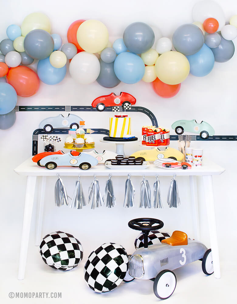 Momo Party Race Car Box, set up inspiration with Meri Meri race car paper plates, race car shaped napkins, race car party paper cups, black strip small plate, neon confetti flag cupcake kit, CandyLab race car wooden toys on top of cake, Fuel treat favor box with popcorns, all on dessert table.  colorful balloon cloud, Road tape with Car plates and Backdrop decorations. Checkerboard foil balloon and Race Car Rider toy for a modern Race car, car themed birthday party
