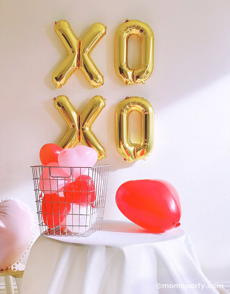 Momo party Valentine's Day box, Party idea with Gold XOXO foil balloons as backdrop wall decoration, Heart Shaped Latex balloons in the silver basket, with a Red heart shaped latex balloon on the table, a pink heart shaped pillow on the side, a small home party set up for a Valentine's Day, Galentine's Day with friends, 