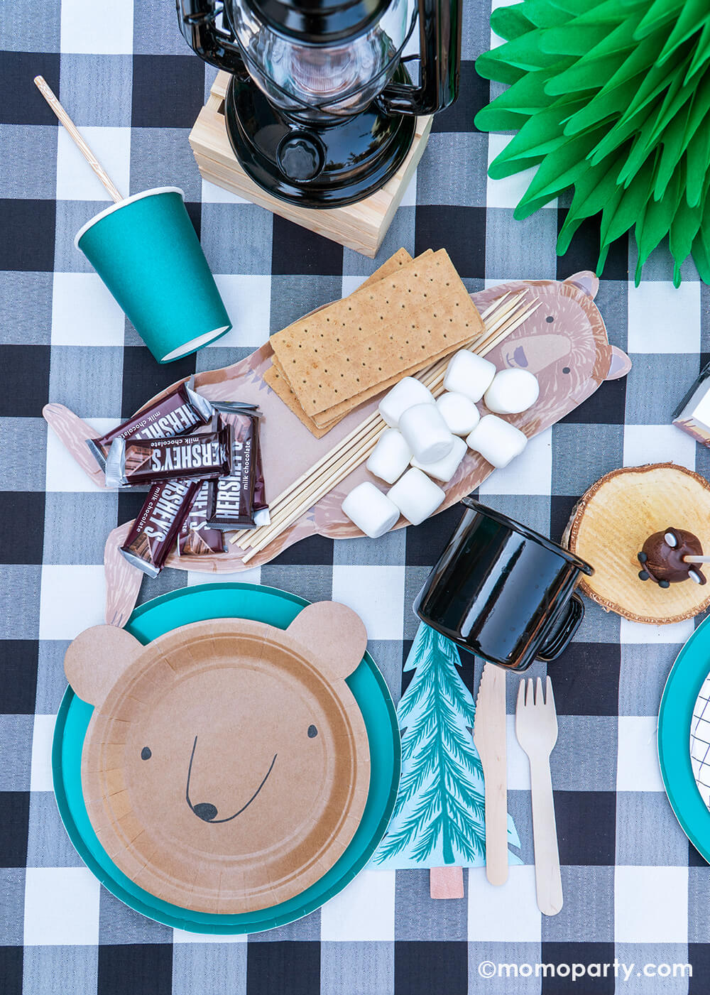Kid's Woodland Camping outdoor birthday party, table top with Bear small plates layered with Oh happy day forest green 9 inch Plates, Meri Meri tree napkins with wooden utensils, black campfire mugs, brown bear platter with Hershey's milk chocolate bar, marshmallows and graham cracker ready for smore, green Tree tissue decor, all over the black and white  Checkered Gingham tablecloth for a modern camping themed birthday party 