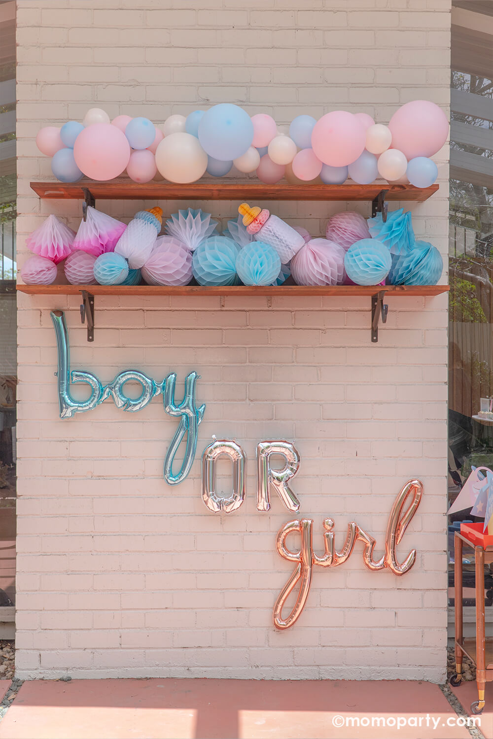Boy or girl? A gender revel party by Momo Party, a brick wall decorated with script foil letter balloons and a wall filled with honeycomb decorations in pink and blue and a boho pastel color balloon garland in baby pink and baby blue colors