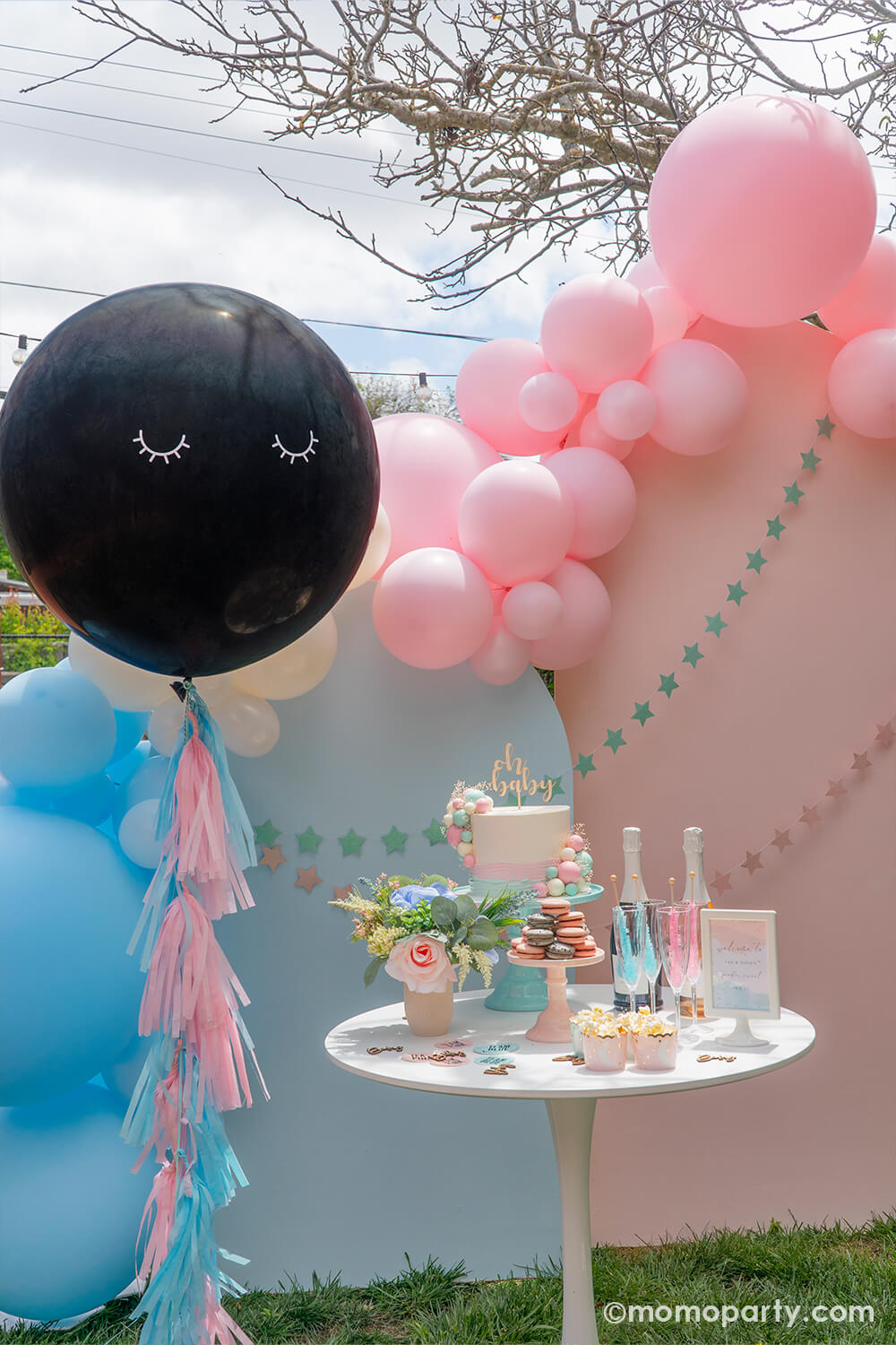 A beautiful gender reveal party set up with pastel blue and pastel pink balloons and a jumbo gender reveal confetti latex decorated with pink and blue tassels and a baby's sleepy eyes, ready to be popped to reveal the baby's gender. On the side is a party table with a gender reveal cake inspired by the balloon garland decoration, topped with oh baby - a darling set up for a modern gender reveal party