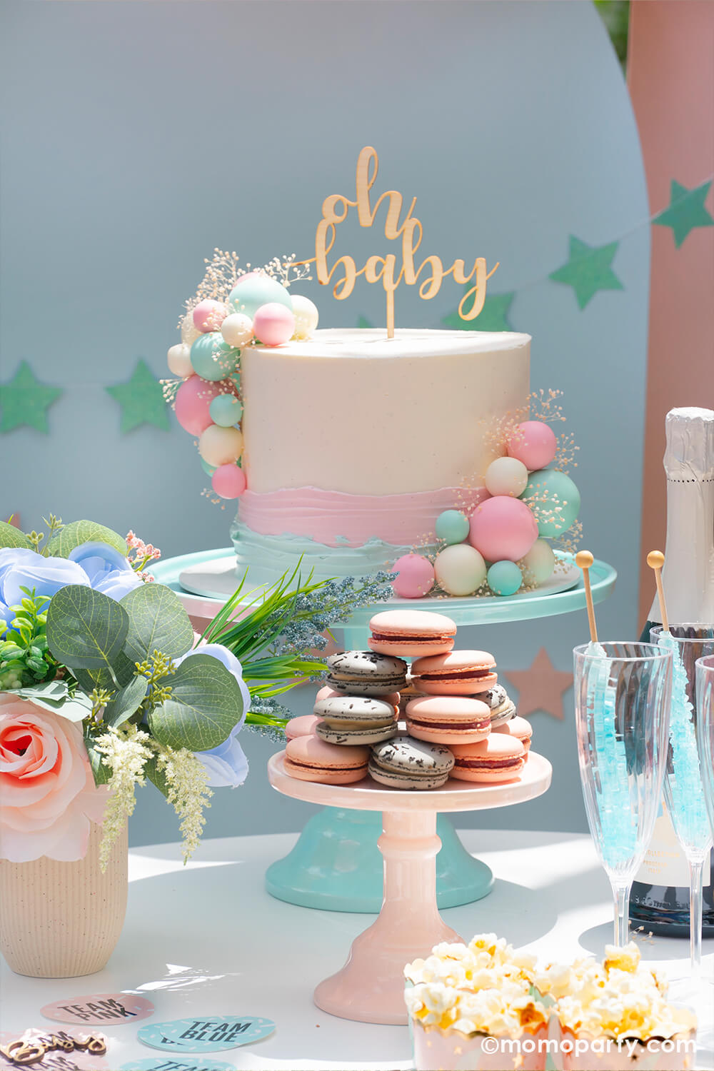 A modern gender reveal cake inspired by an organic balloon arch designs topped with Party Deco's wooden oh baby cake topper, on the side is a pile of blue and pink french macaroon and bubbly drinks in pink and blue, perfect inspiration for a modern baby gender reveal party