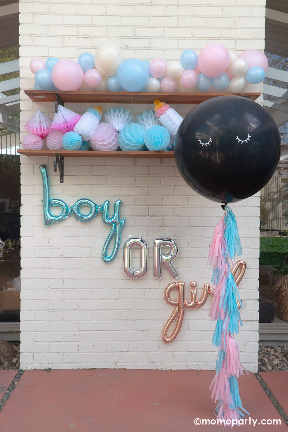 Boy or girl? A gender revel party by Momo Party, a brick wall decorated with script foil letter balloons and a wall filled with honeycomb decorations in pink and blue and a boho pastel color balloon garland in baby pink and baby blue colors. In the front is the jumbo gender reveal confetti balloon decorated with pink and blue tassel, ready to be popped to reveal baby's gender!