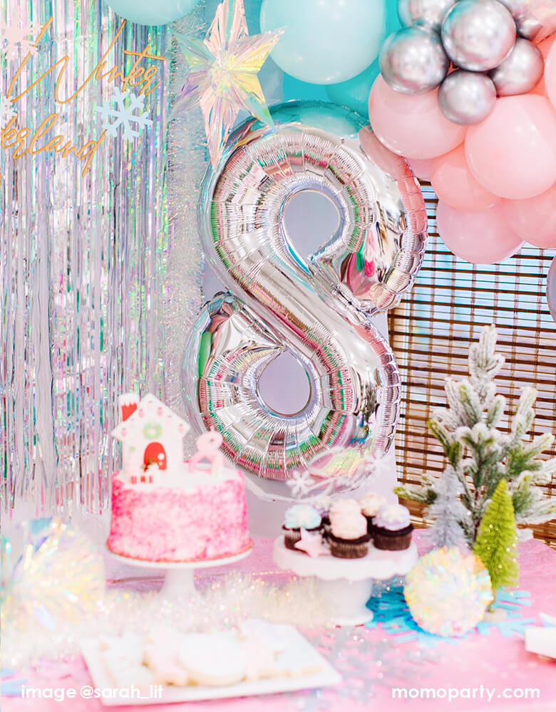 a Dreamy Winter Wonderland themed 8th years birthday Party set up with large number 8 silver foil balloon, in front of iridescent curtain, with iridescent hanging starts, desserts and cake on top of pink table cloth with christmas tree decorations  