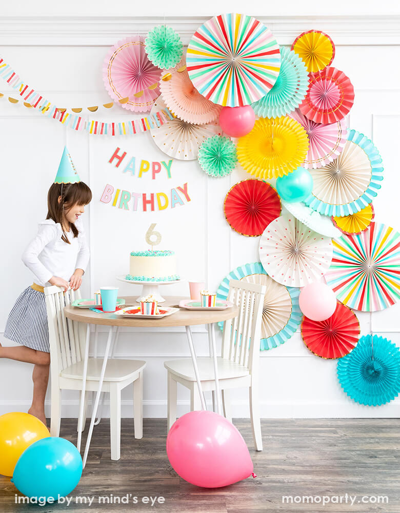 A happy girl wearing a blue paper party hat and standing next to a buttercream cake with gold number six cake topper on it at her 6 years old birthday party. there are colorful paper tablewares on the table, a colorful Happy Birthday letter Banner on the wall, and My mind's eye Hip Hip Hooray Paper Fans filled the whole wall for a bright and cheery backdrop, colorful balloons on the ground. This colorful party collection are perfect for summertime ice cream party, pool party to bright birthday bashes