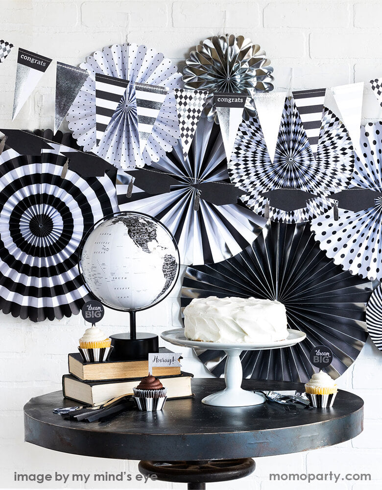 A modern black and white Graduation Party, decorated with My Minds Eye Graduation Party Fan layered with graduation hat Banner on top. a white cake on the cake stand, books, black and white globe as decorations, cupcakes with Graduation cupcakes kit around the table. Celebrate your Class of 2021 grad with this modern graduation collection.