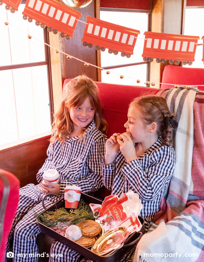 Two little girls in their Holiday pajamas enjoying a vintage tin tray filled with holly branches, holiday treats and cookies and Momo Party's North Pole Express train shaped plates, bell shaped side plates and polar express red 8 oz to-go cups by My Mind's Eye sitting on a vintage Polar Express train cabin decorated with Momo Party's North Pole Express train and bell garland set, a perfect idea for this Holiday season's celebration with family and kids.