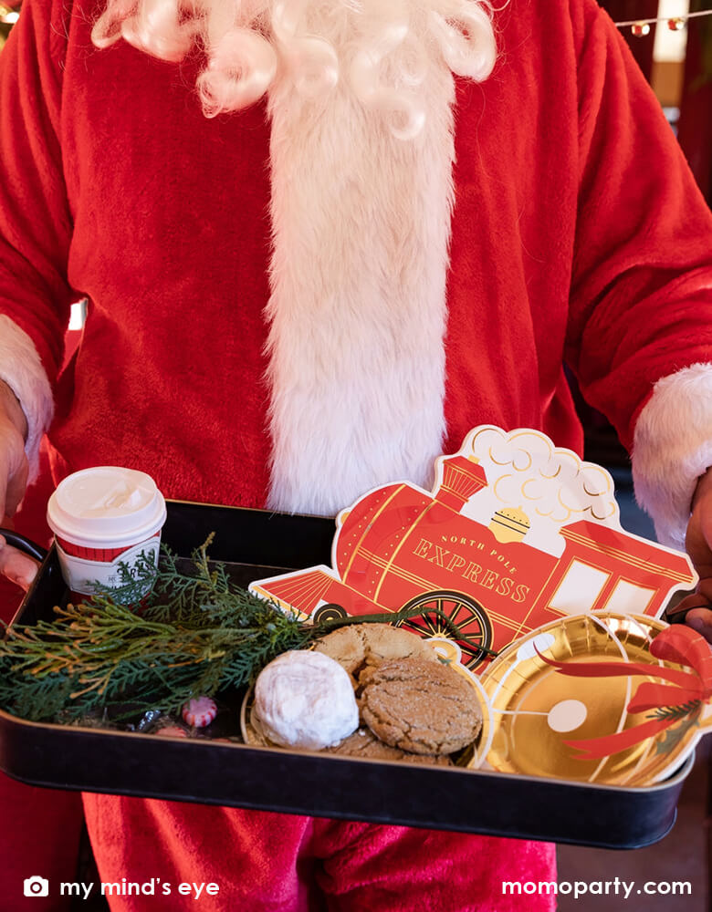Santa holding a vintage tin tray filled with holly branches, holiday treats and cookies and Momo Party's North Pole Express train shaped plates, bell shaped side plates and polar express red 8 oz to-go cups by My Mind's Eye on a vintage Polar Express train cabin red leather seat, ready for a festive Holiday celebration for family and kids.
