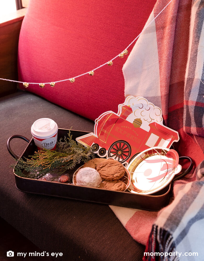 A vintage tin tray filled with holly branches, holiday treats and cookies and Momo Party's North Pole Express train shaped plates, bell shaped side plates and polar express red 8 oz to-go cups by My Mind's Eye on a vintage Polar Express train cabin red leather seat, ready for a festive Holiday celebration for family and kids.