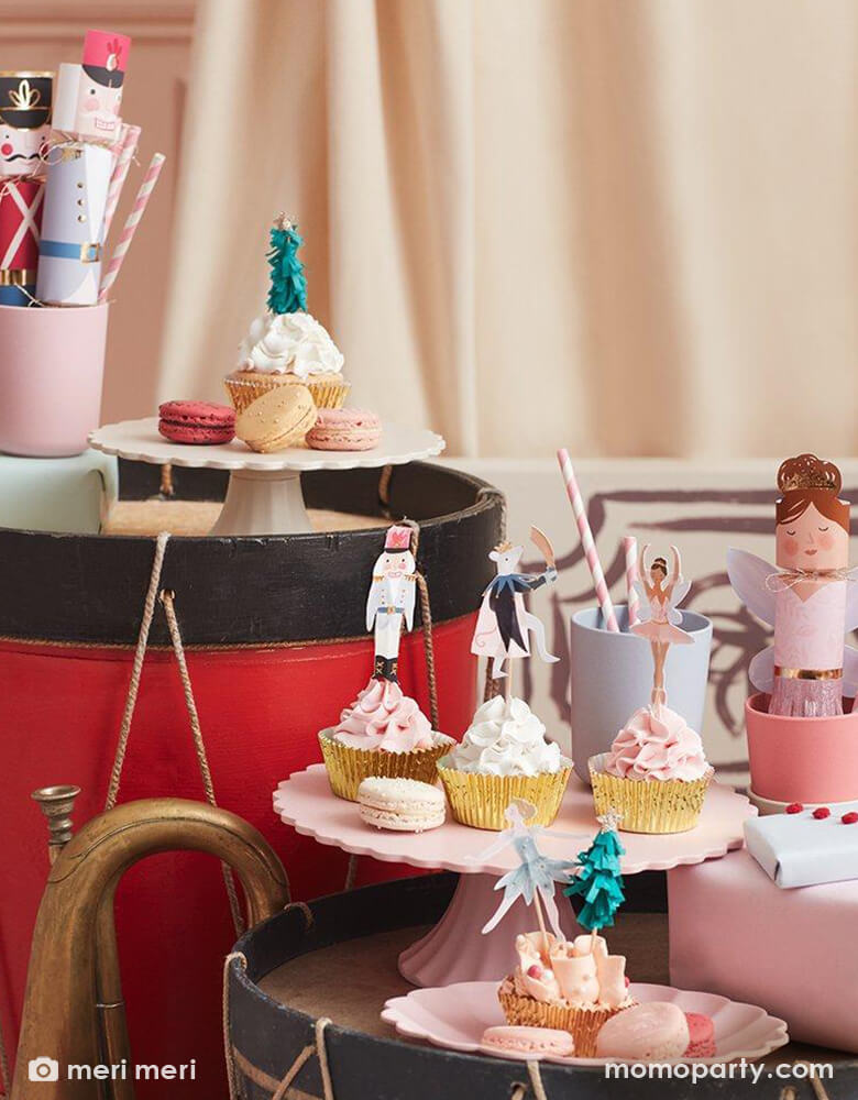 Cupcakes decorated with Nutcracker themed cupcake toppers from Meri Meri Nutcracker Cupcake Kit. With opper designs include Nutcracker characters and a green fringed crepe paper Christmas tree with gold foil star on top. With gold foil cupcake cases on a pink and white cake stands. There are Nutcracker Character Party Crackers in the cups, These modern holiday party supplies  make this Christmas celebration super special.