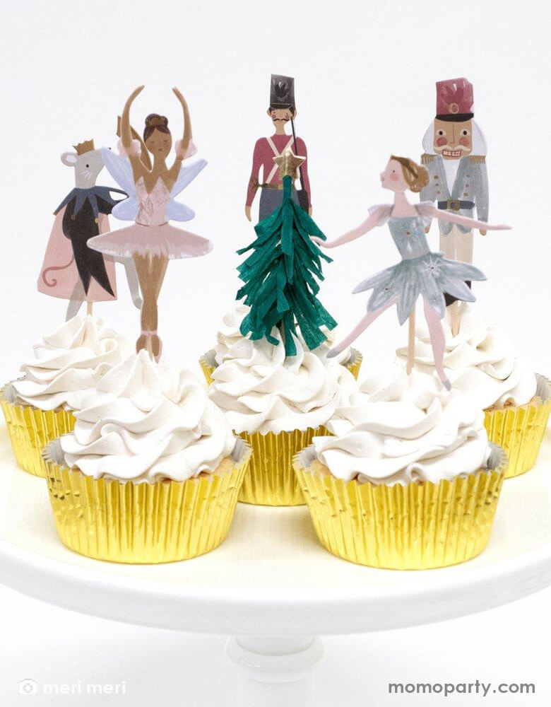 White Cupcakes decorated with Nutcracker themed cupcake toppers from Meri Meri Nutcracker Cupcake Kit. With opper designs include Nutcracker characters and a green fringed crepe paper Christmas tree with gold foil star on top. With gold foil cupcake cases on a pink cake stand.  They make these cupcakes look super special this Christmas celebration.