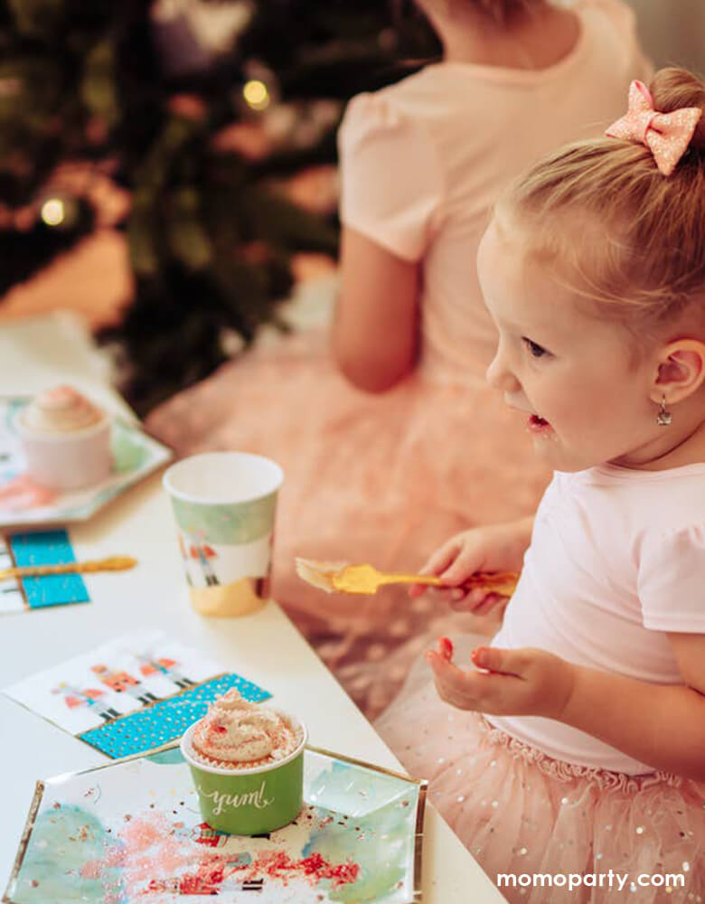 little girl wearing a pink fairytale tutu dress with pink glitter bow tie hair piece, happily eating a cupcake, with Nutcracker Holiday Party Plates and napkins on the table, which printed with whimsical classic Nutcracker characters of Nutcracker, Mouse King designs, sitting around her family and friends in a warm cozy Holiday dinner table. these high quality eco friendly party cups look so on theme for your home or a nutcracker lover christmas party