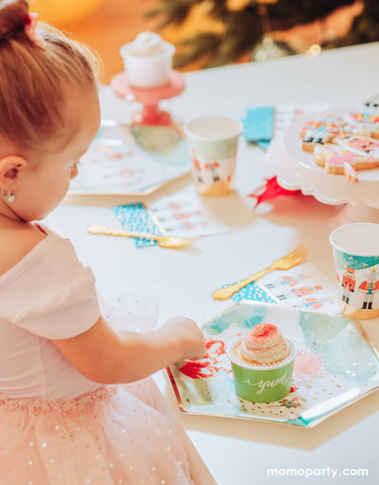 a little girl wearing a pink fairytale tutu dress with pink glitter bow tie hair piece, looking at a cupcake on a warm cozy Holiday dinner table, setted up with Nutcracker Holiday Party Plates, napkins and cups, cookies on the cake stand. these paper partyware printed with whimsical classic Nutcracker characters of Nutcracker, Mouse King designs, these high quality eco friendly party cups look so on theme for your home or a nutcracker lover christmas party