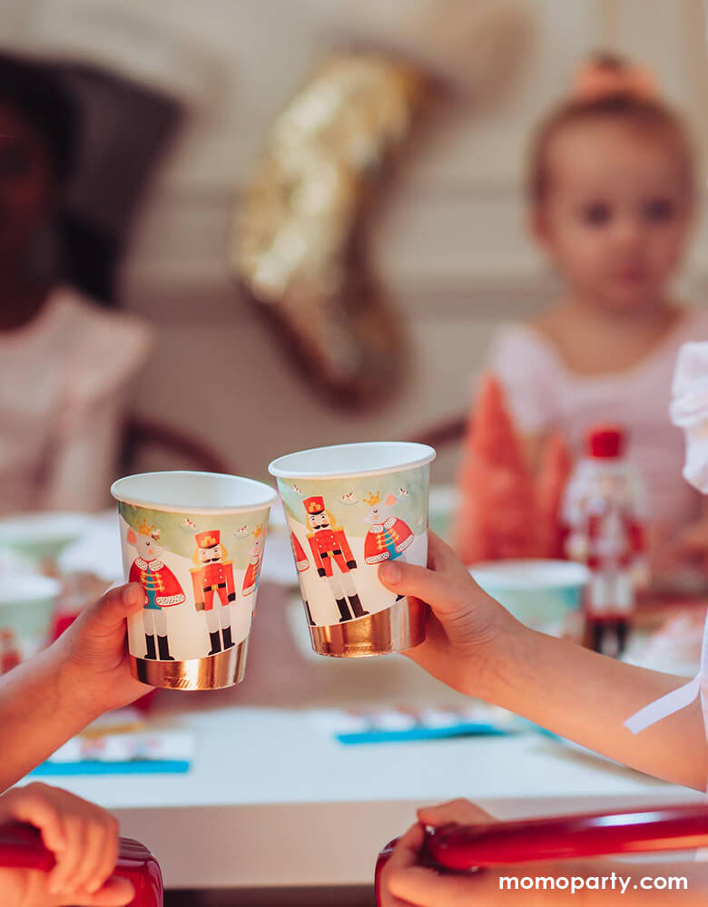  on a warm cozy Holiday dinner table, the little hands cheers with holding Nutcracker Holiday Party cups, there cookies on the cake stand. little friends sitting around, these paper cups printed with whimsical classic Nutcracker characters of Nutcracker, Mouse King designs, these high quality eco friendly party cups look so on theme for your home or a nutcracker lover christmas party