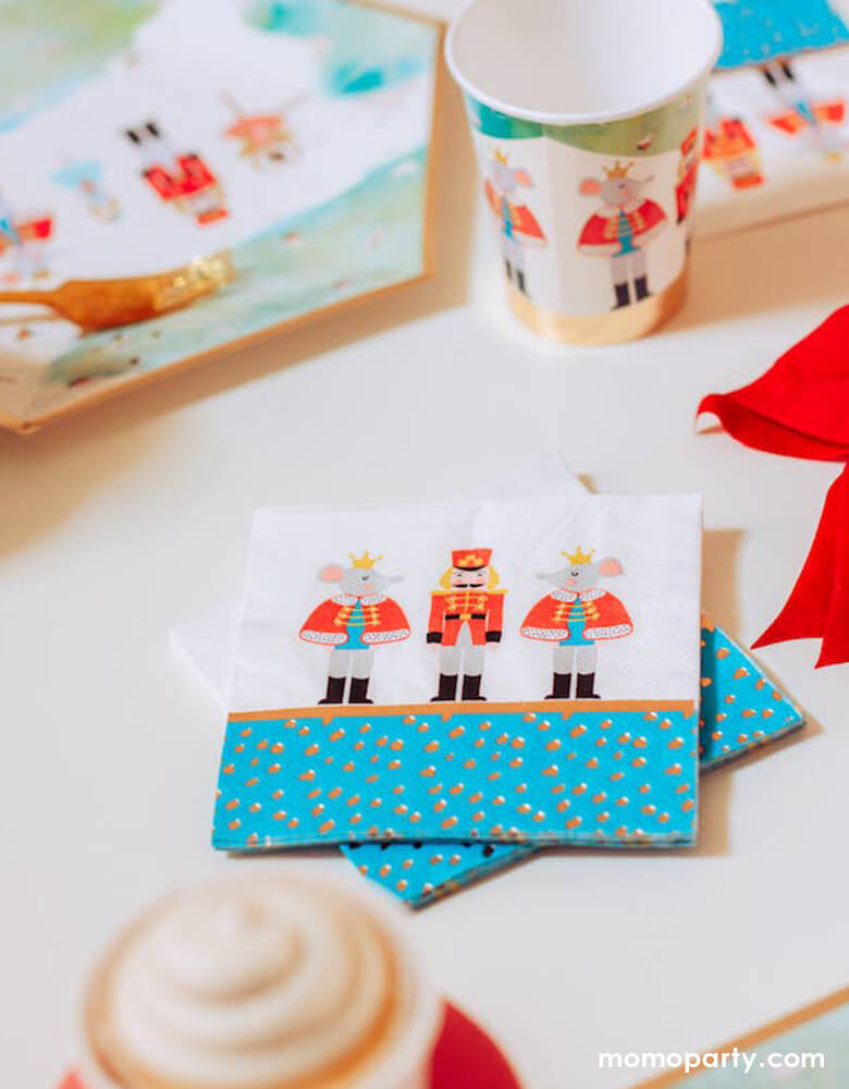 Holiday party table close up of Crated party Nutcracker Holiday Party Napkins, with whimsical classic Nutcracker characters, like Nutcracker, Mouse King designs, and nutcracker holiday party cups and plates, red ribbon, gold folks around. Adding these well designed party supplies to set a scene worthy of a standing ovation for your home or a nutcracker lover christmas party