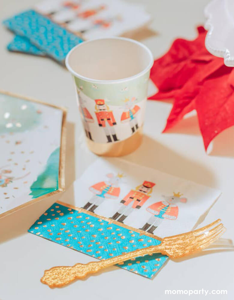 Holiday party table close up of Crated party Nutcracker Holiday Party Napkins, with whimsical classic Nutcracker characters, like Nutcracker, Mouse King designs, and nutcracker holiday party cups and plates, red ribbon, gold folks around. Adding these well designed party supplies to set a scene worthy of a standing ovation for your home or a nutcracker lover christmas party