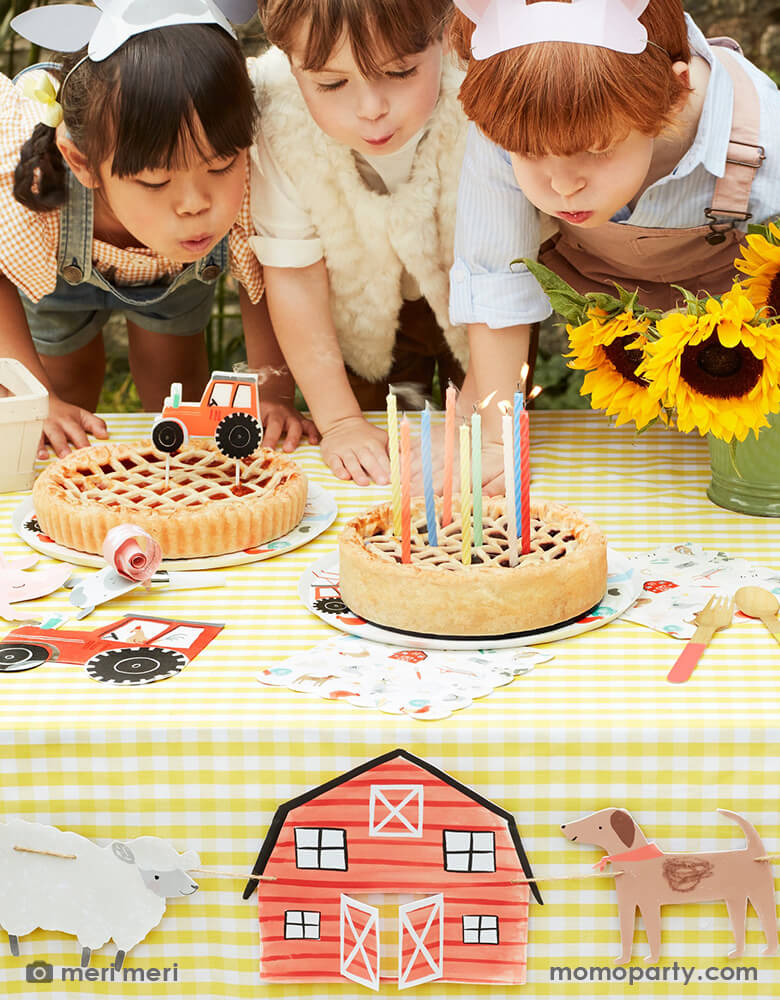 3 Kids wearing On The Farm Animal Ears on their head, blowing candles on the pie, at a Farm themed birthday party table. This fun modern fun look table filled with  Apple Pie with On the Farm Tractor Candle, sunflowers, On The Farm Tractor Napkins, On The Farm Large Napkins on a light yellow plaid tablecloth, with a On-The-Farm-Large-Garland hanging in the front of the table.
