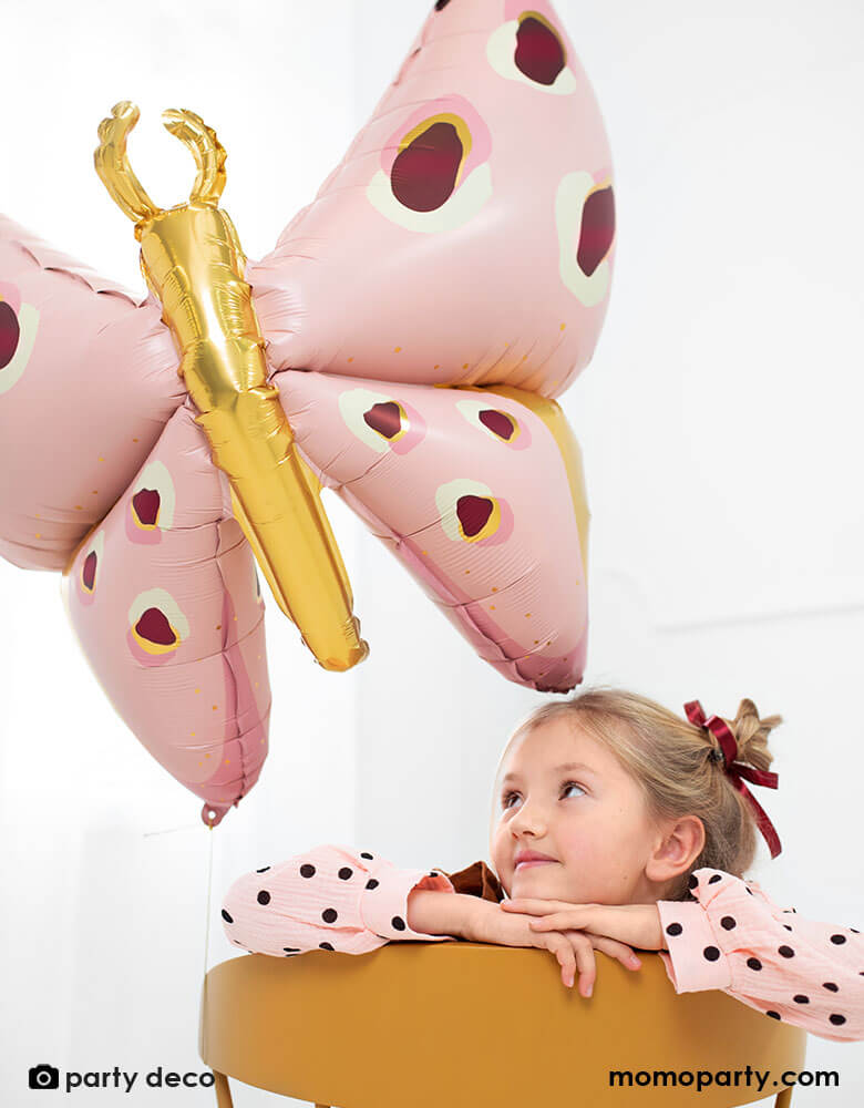 A little girl sitting on a wooden chair looking at Party Deco's 47" Beautiful Pink Butterfly Foil Balloon in pretty pastel pink and maroon with gold accent in the back next to a white window, creates a whimsical dreamy scene