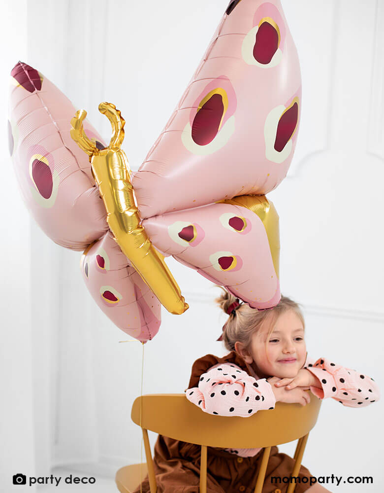 A little girl sitting on a wooden chair smiling looks as she's daydreaming, with Party Deco's 47" Beautiful Pink Butterfly Foil Balloon in pretty pastel pink and maroon with gold accent in the back next to a white window, creates a whimsical dreamy scene