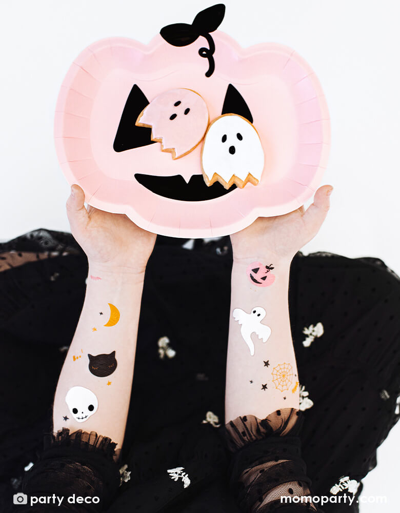 A girl in her black Halloween costume with her arms decorated with Party Deco's Boo! Halloween temporary tattoos featuring not-so-spooky Halloween characters including black cats, skulls, moon, ghosts, and bats, holding a pink pumpkin shaped plates with ghost shaped sugar cookies on it
