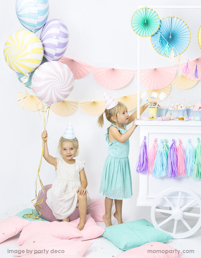 A kid wearing a party hat and holding Party Deco pastel candy foil balloons A sweets themed summer party. Next to her there is a girl getting snake from dessert cart filled with pastel decorations and mashmaros, candies.