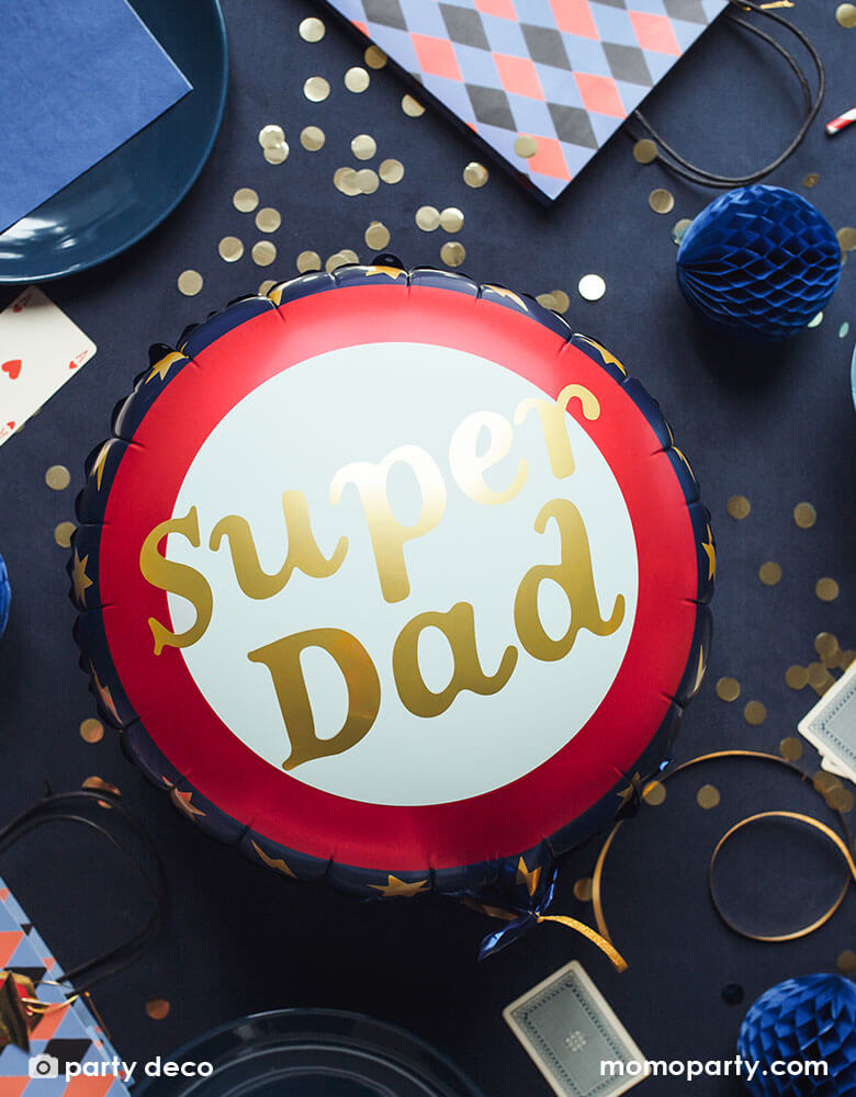 Party Deco 18" Round blue, red and gold foil balloon with Super Dad message on it on a party table filled gold confetti, blue honeycomb balls, cards, and gift bags for a special Father's Day celebration