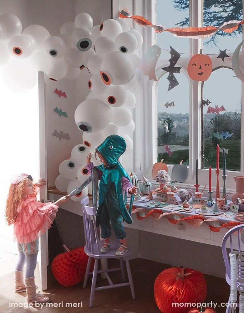 A not-so-spooky Halloween party decoration set up by Meri Meri featuring Momo Party's pastel Halloween party supplies including an eyeball balloon garland, pastel Halloween stitched streamer and pastel Halloween large garland hung on the wall. On the table was set with various pastel Halloween themed tableware including pumpkin shaped plates, napkins and party cups. In the front two kids dressed up in costume having fun at the party.