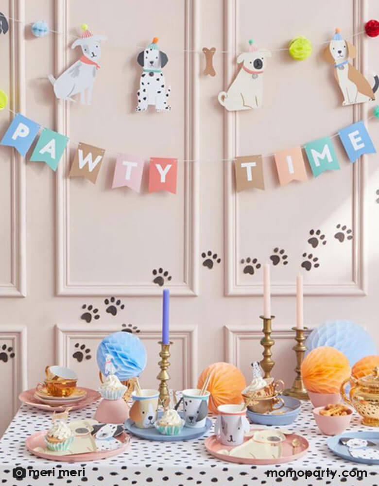 An adorable party set up for puppy dog themed birthday, featuring Momo Party's 6 ft Pawty Time party garland by Meri Meri, it features furry friends with embellished tails and pompom hats, bones and honeycomb balls. Drape it on the party table or on a wall for a delightful decoration. It's perfect for a dog lover's birthday party or a party for your family pet. In the front, a party table filled with puppy themed party supplies including dog party cups, pug shaped plates and Dalmatian shaped napkins.