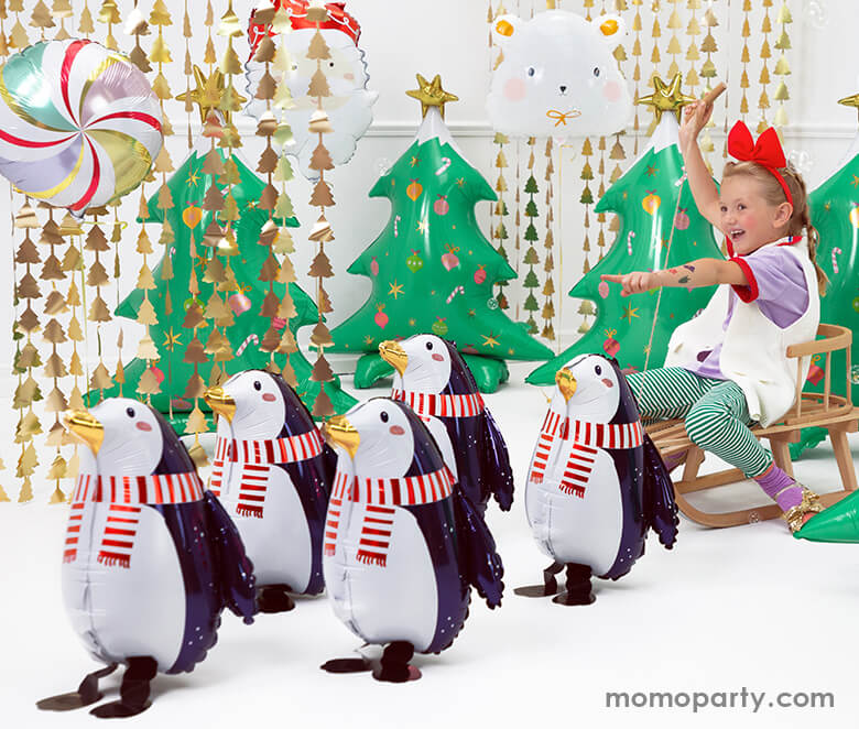Christmas winter wonderland celebration with lots of Penguin Shaped Foil Mylar Balloon walking in from of Christmas Tree Gold Backdrop, Christmas Tree shaped foil balloon and a girl sitting on a snow slider