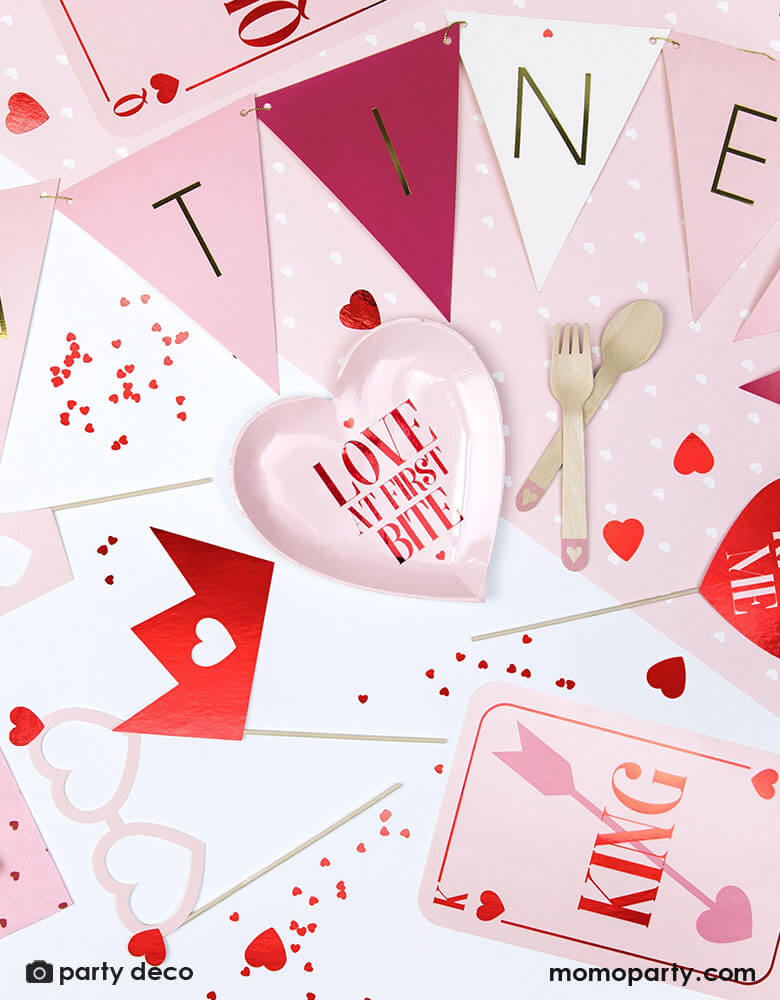 A table of Valentine's Day themed party supplies by Party Deco including heart shaped plates, pink party banner, red heart shaped confetti, photobooth props and pink cutlery with heart design on it. 