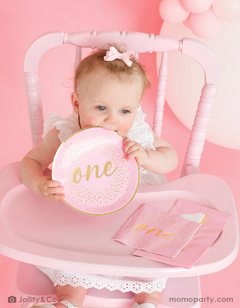 A baby girl in cute pink bow sitting on a vintage pink highchair holding Jollity 7-inch onederland pink dessert plate with number 1 and botanical boho greenery illustrations in beautiful gold foil along with Jollity's onederful pink napkin and guest towel spread on the highchair tray, celebrating her 1st birthday party with baby pink balloon garland in the backdrop