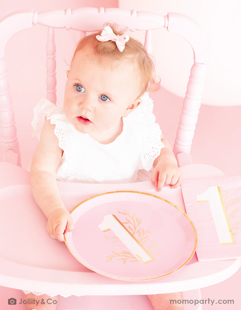 A baby girl in cute pink bow sitting on a vintage pink highchair holding Jollity 9-inch pink round dinner plate with number 1 and botanical boho greenery illustrations in beautiful gold foil for celebrating baby's 1st birthday party