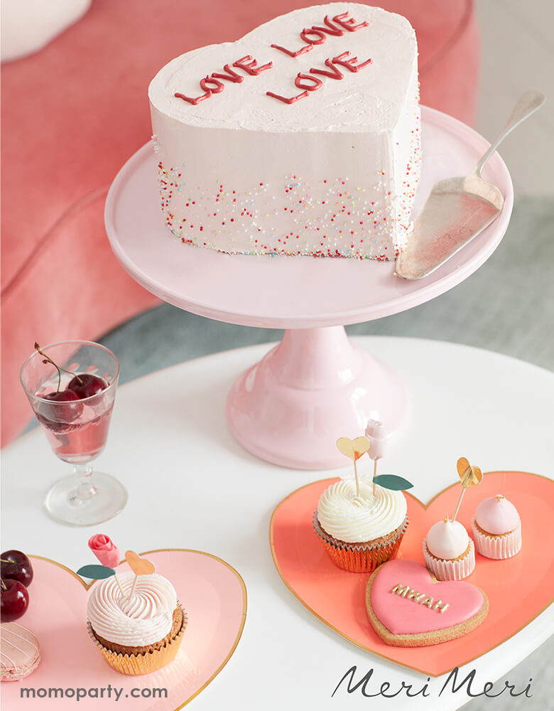 A Valentine's Day themed dessert table with a heart shaped buttercream cake decorated with the message of "love love love" along with Meri Meri's pink tone heart shaped plates filled with Valentine themed treats including heart shaped sugar cookies and cupcakes decorated with floral and heart shaped toppers, and cherry infused drinks